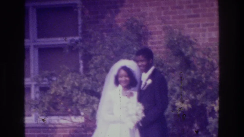 Home-movie frame from the Bronx in 1975 showing newlyweds close together outside a brick building.