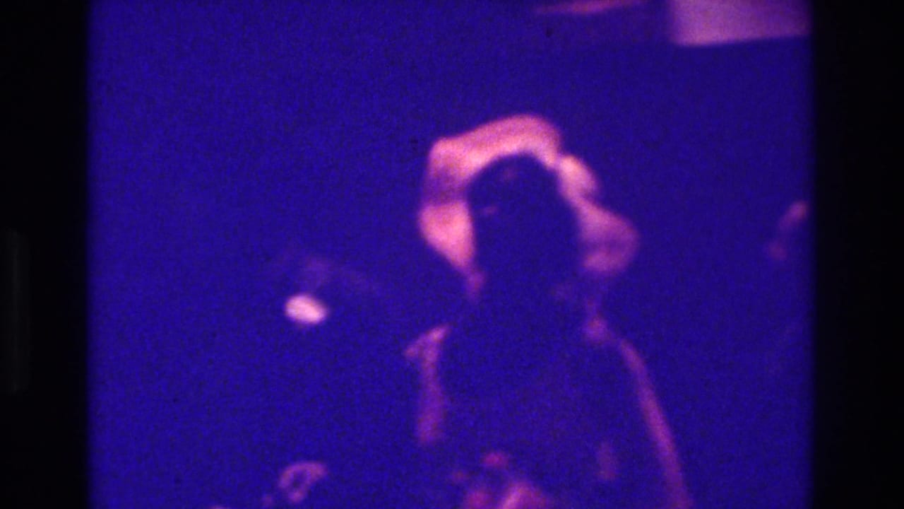 1975 Bronx 8mm home-movie frame showing a blurred woman in light clothing among darker figures at an indoor celebration.