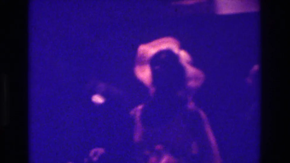 1975 Bronx 8mm home-movie frame showing a blurred woman in light clothing among darker figures at an indoor celebration.
