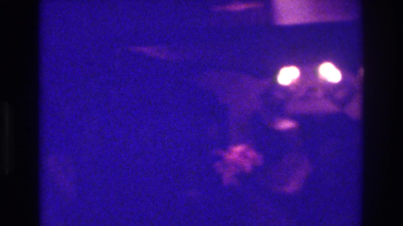 Dark, grainy frame with bright lights and faint figures in a low-lit interior.
