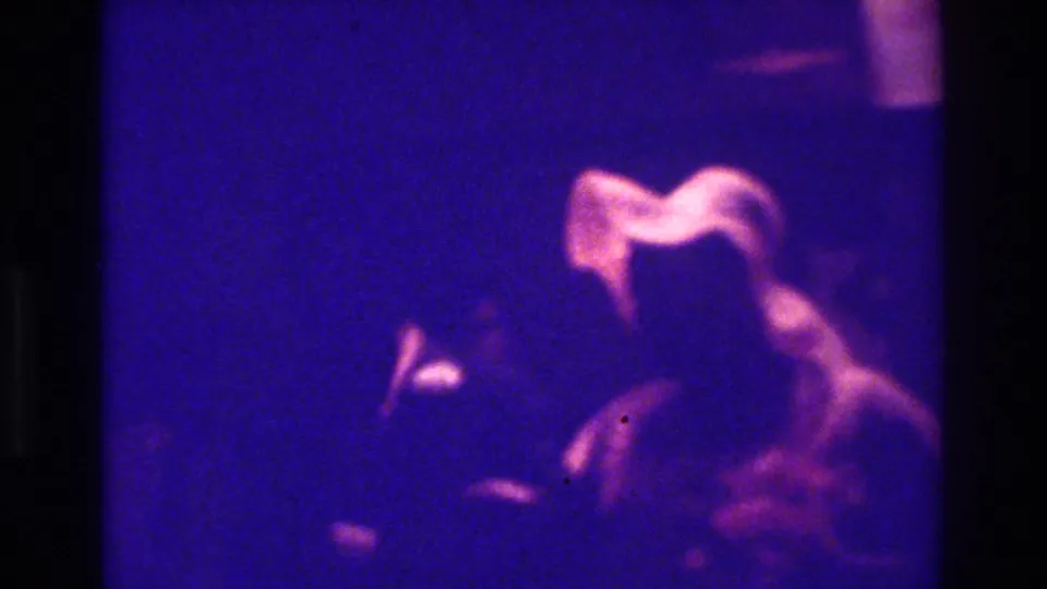 1975 Bronx 8mm frame of a blurred woman in light clothing beside a dark-clothed figure during an indoor celebration.