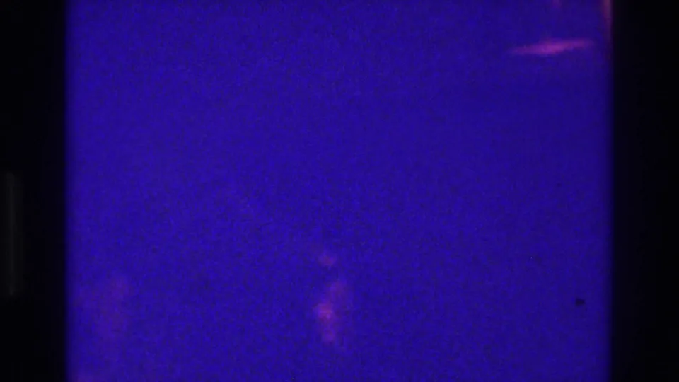Very dark 1975 Bronx 8mm home-movie frame with faint guest shapes in a low-light indoor celebration.