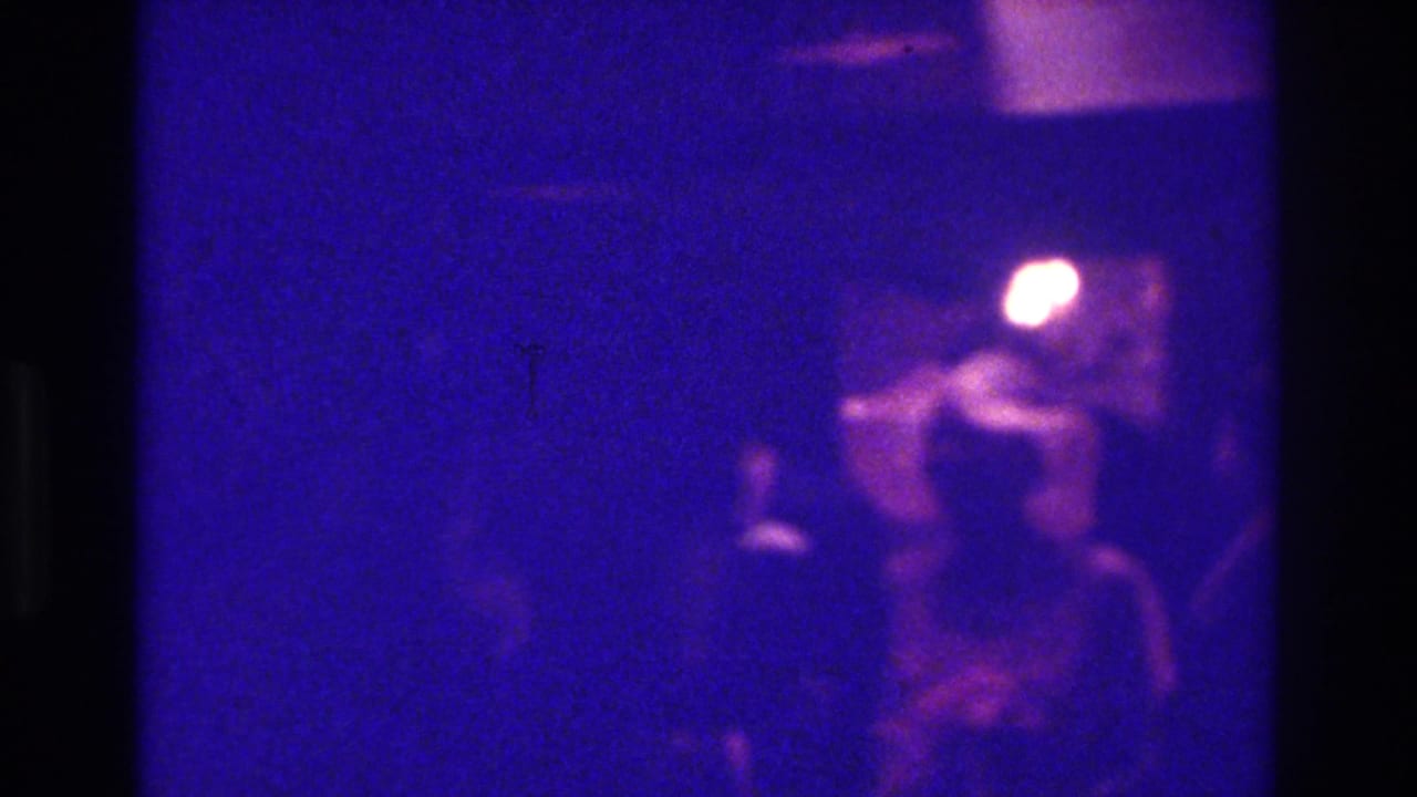 Dark indoor scene with a seated or standing figure near glowing lights.