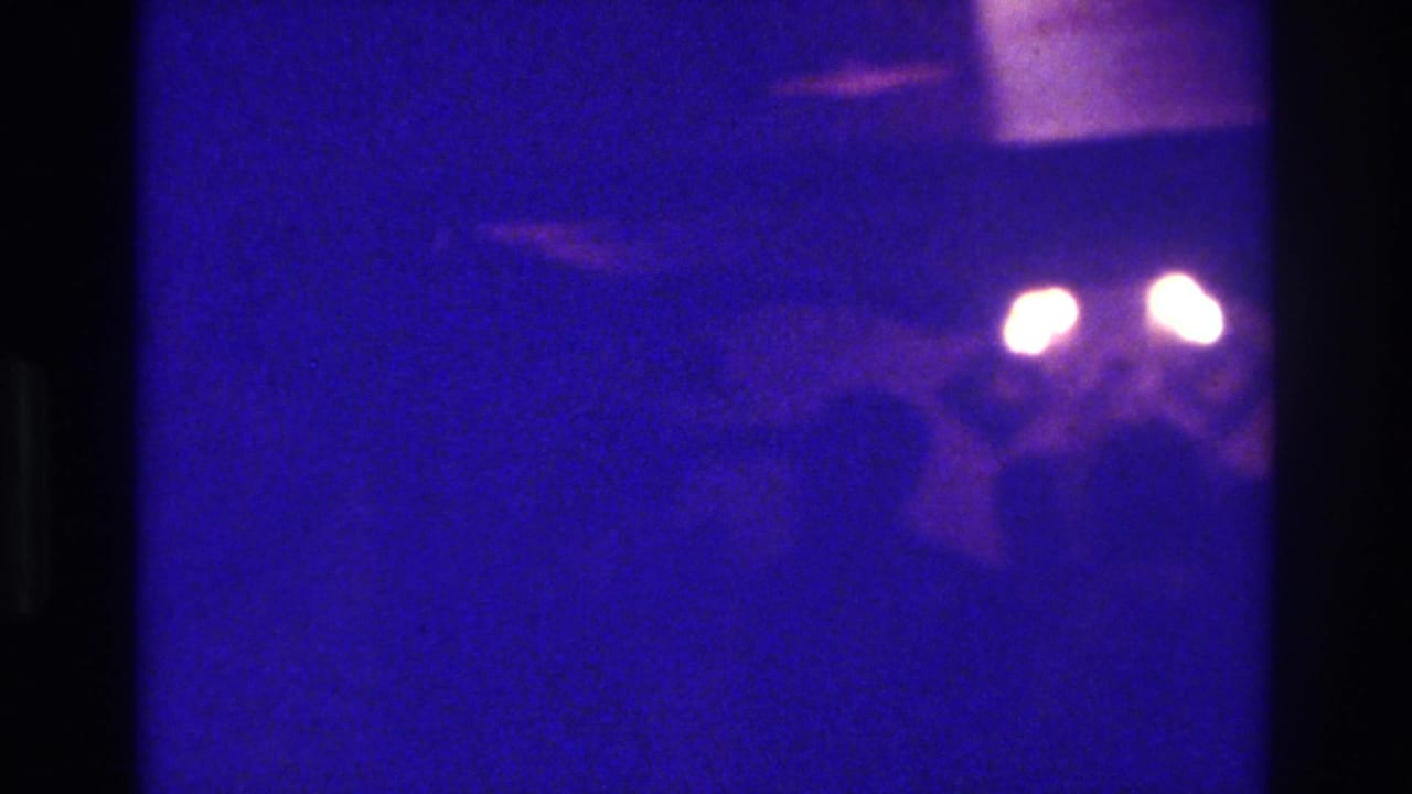1975 Bronx 8mm frame with blue-purple darkness, grain, indistinct figures, and two glowing lights in soft focus.