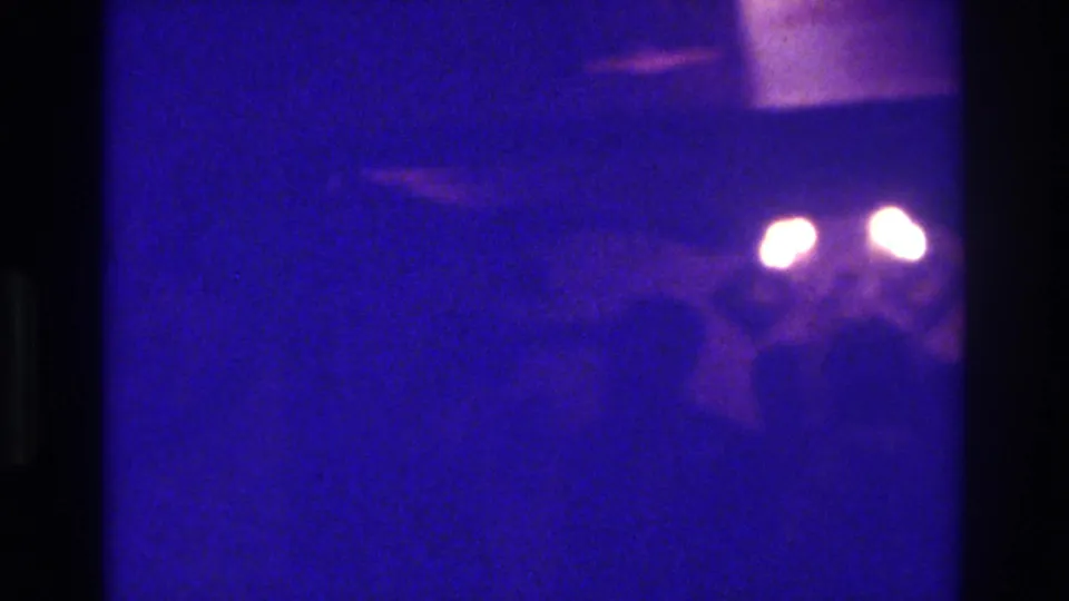 1975 Bronx 8mm frame with blue-purple darkness, grain, indistinct figures, and two glowing lights in soft focus.