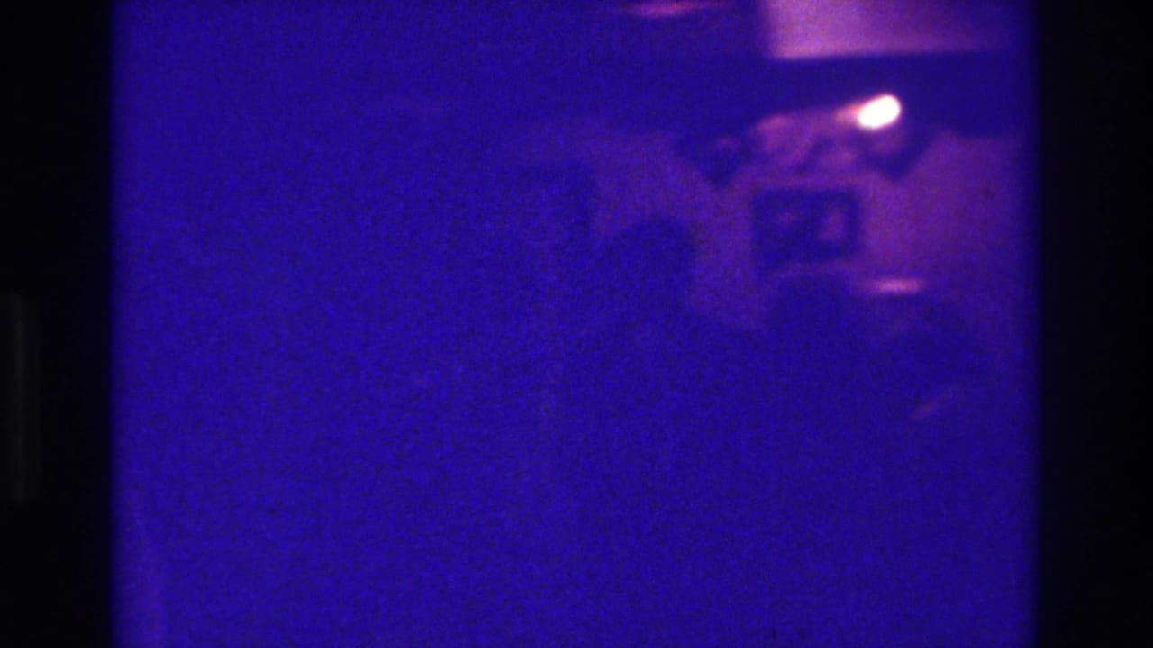 Dim blue-purple frame with a bright camera flash near the top and indistinct people below.