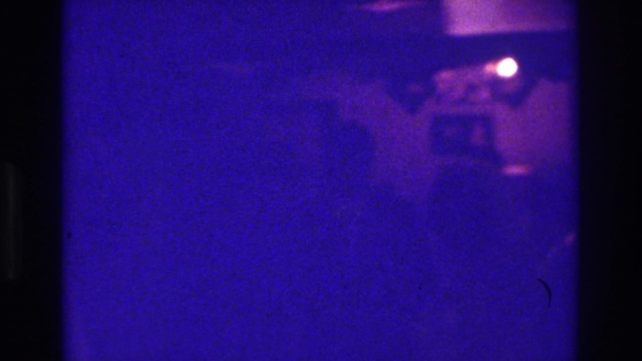 A dim 1975 Bronx wedding frame shows guests in shadow under strong blue-purple film color shift and bright flash reflections.