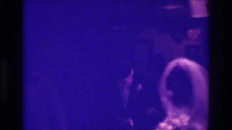 In the 1975 Bronx wedding footage, a bride in a white veil moves through a dark indoor crowd under blue-purple 8mm color distortion.