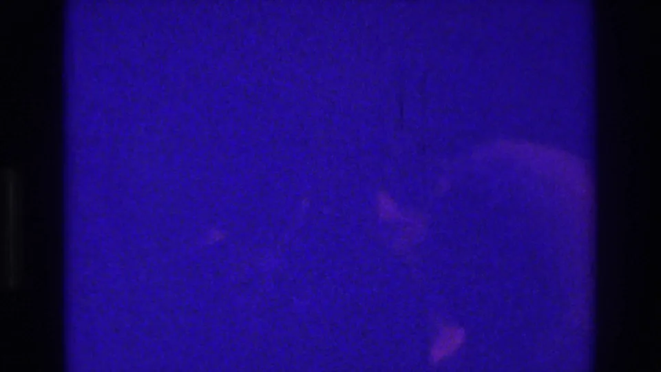 1975 Bronx 8mm frame showing a dim blue-purple space, dark vertical obstructions, soft light reflections, and heavy film grain.