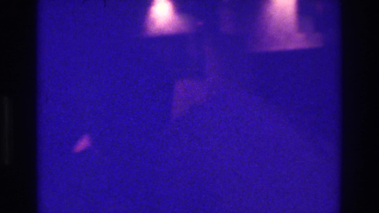 1975 Bronx 8mm frame with a central dark obstruction, blue-purple cast, faint warm highlights, and indistinct background shapes.