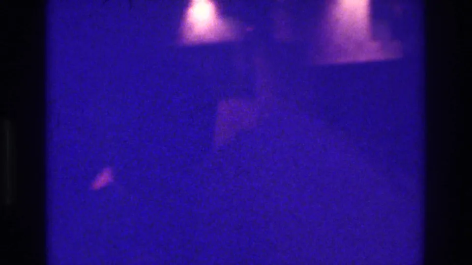1975 Bronx 8mm frame with a central dark obstruction, blue-purple cast, faint warm highlights, and indistinct background shapes.