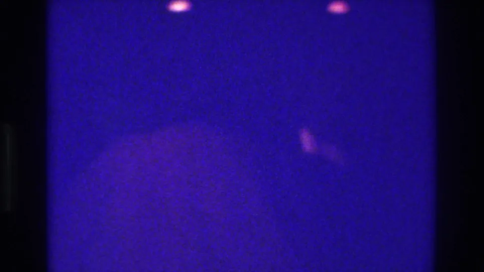 1975 Bronx 8mm frame showing grainy blue-purple illumination, dark frame edges, faint light spots, and no identifiable people or signage.
