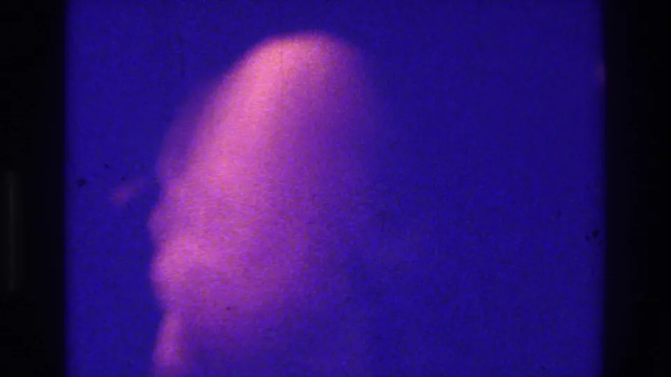 1975 Bronx 8mm frame with an indistinct pale figure or sheeted form emerging in blue light, with heavy grain and soft focus.