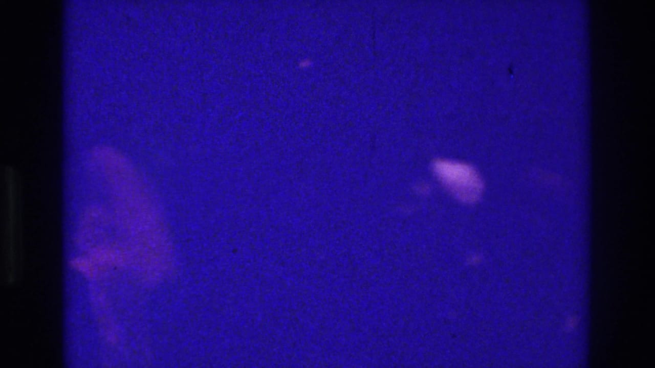 1975 Bronx home-movie frame with a dim blue-purple pub interior and indistinct shadowed figures or reflections.