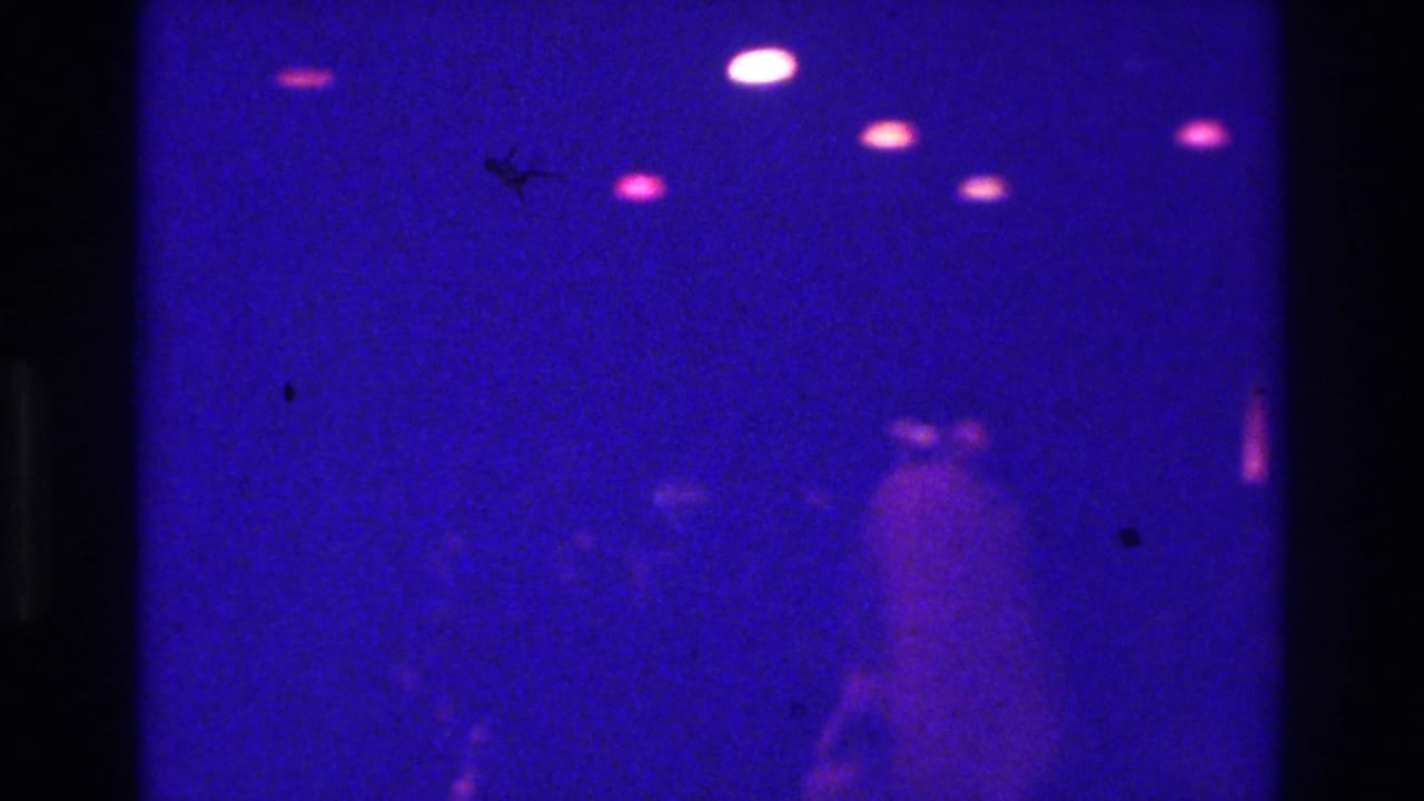 1975 Bronx nightclub home-movie frame with faint silhouettes moving under blue-purple light and bright ceiling flares.