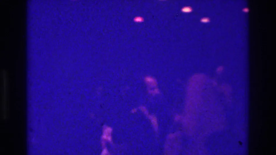 1975 Bronx nightclub frame with blurred silhouettes clustered low in frame under dim blue-purple lighting.