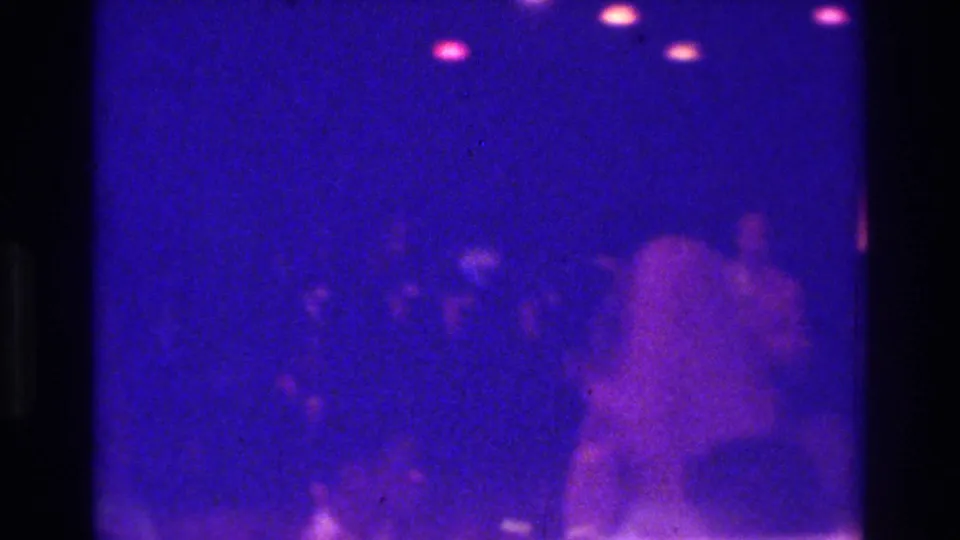 1975 Bronx nightclub home-movie frame with multiple soft silhouettes and pink light flares in a blue-lit interior.