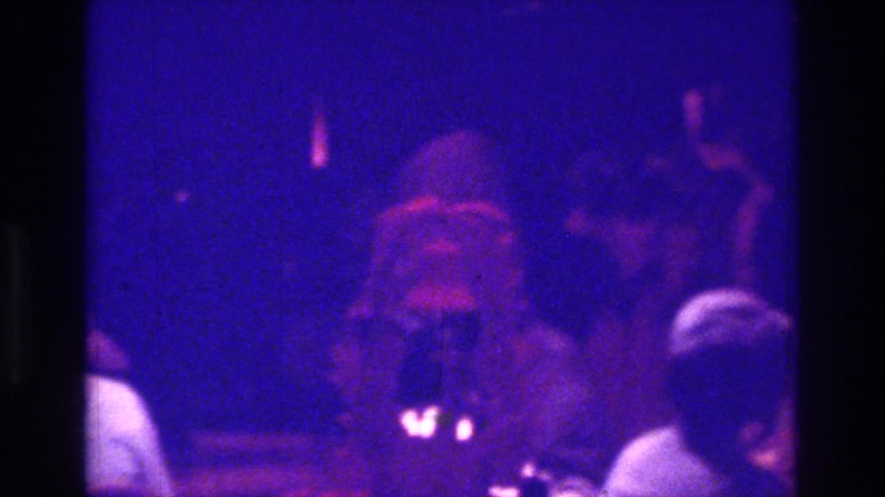 1975 Bronx 8mm wedding reception frame with blue-purple lighting, foreground guests, and a soft central figure in light clothing.