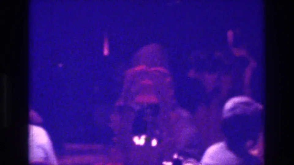 1975 Bronx 8mm wedding reception frame with blue-purple lighting, foreground guests, and a soft central figure in light clothing.