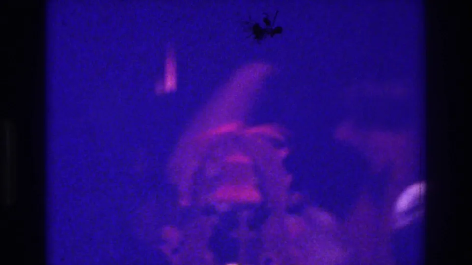 1975 Bronx 8mm reception frame with a closer, blurred view of the light-dressed figure amid dark foreground silhouettes and colored lighting.