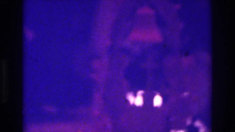 1975 Bronx wedding reception frame with heavy grain, deep blue-purple light, a blurred central figure, and small bright highlights near the lower right.