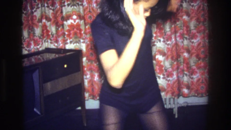 The dancer stands in the living room with one hand near her face, framed by floral curtains and a wood-cased television.