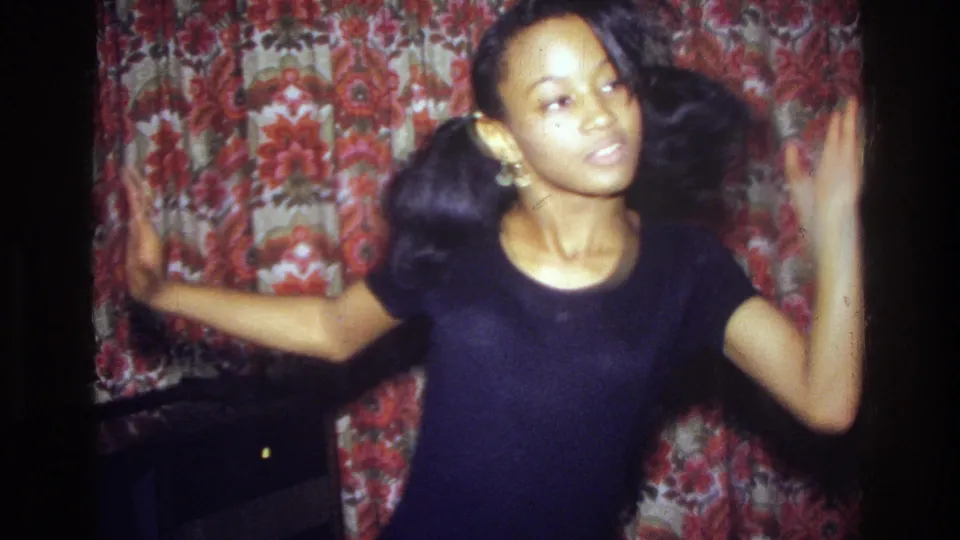 A dancer faces the camera with arms raised, pigtails and earrings visible against floral curtains in the 1975 Bronx living room.