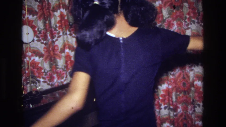 In 1975 Bronx home-movie footage, a woman in a black dress moves near red floral wallpaper and dark furniture inside a private room.