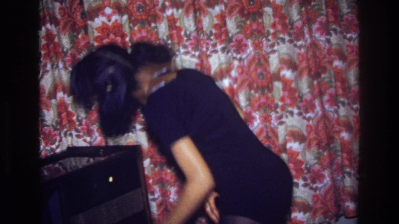 The woman leans forward mid-dance in a dim Bronx interior, with red floral wall covering behind her and a dark cabinet at her side.
