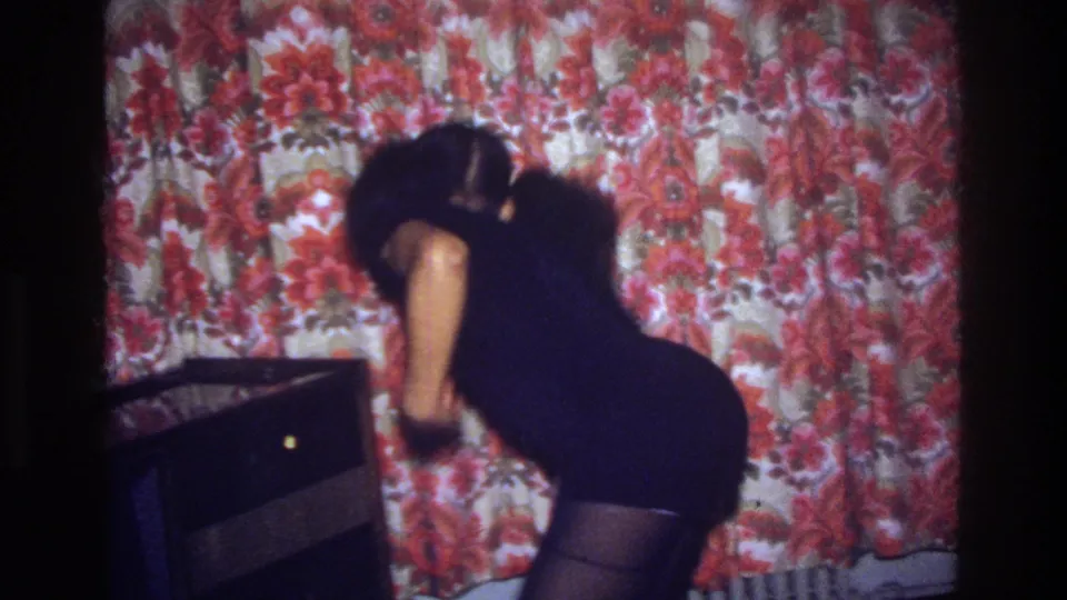 A 1975 home-movie frame shows the woman dancing low near a dark furniture piece, with saturated red floral wallpaper filling the room.