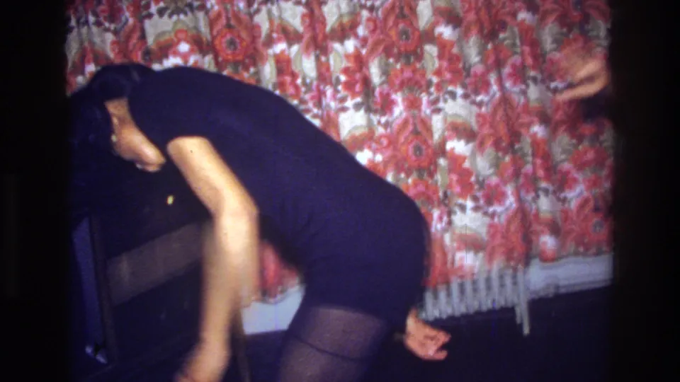The dancer leans sideways across the frame in the Bronx interior, with dark tights, a black dress, and red floral wall covering visible.