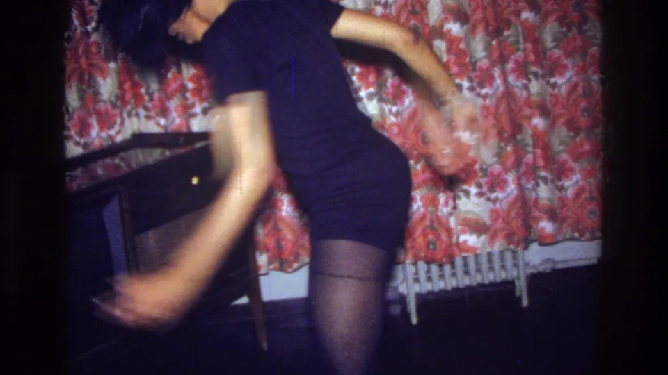 In a 1975 Bronx home-movie frame, a woman in a black dress bends while dancing beside a red floral curtain and dark furniture.