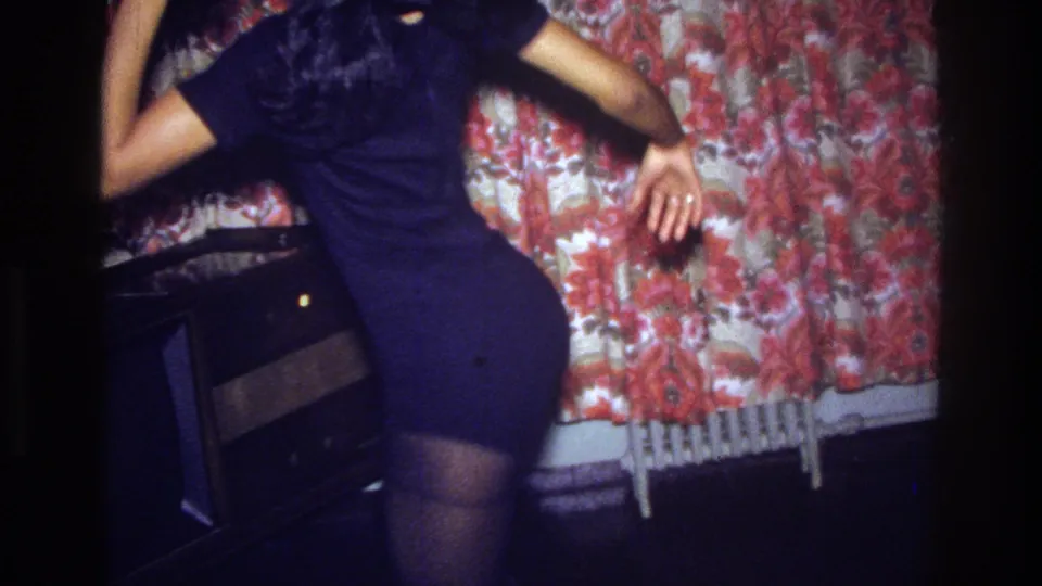 A 1975 Bronx home-movie frame shows the woman dancing in profile near a floral curtain, with a radiator partly visible below.