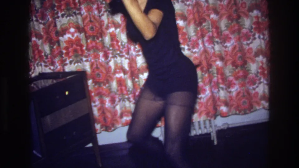 The woman continues dancing in a 1975 Bronx interior, partly cropped by the frame with floral curtain, radiator, and dark furniture visible.