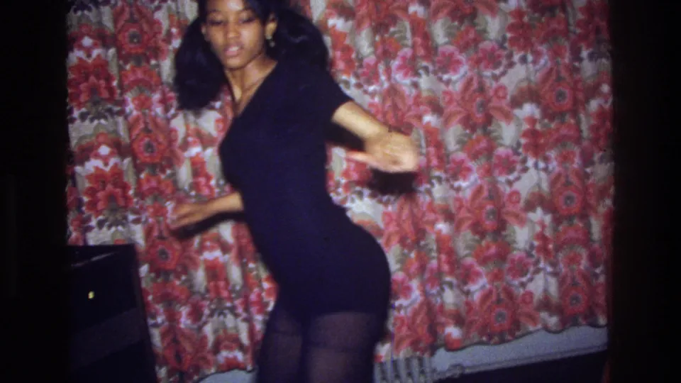 A 1975 Bronx home-movie frame shows the woman facing the camera mid-dance, with floral curtain, radiator, and dark furniture around her.