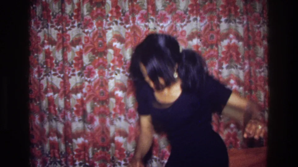 In a 1975 Bronx home-movie frame, a woman in a black dress leans into a dance move before red floral curtains.