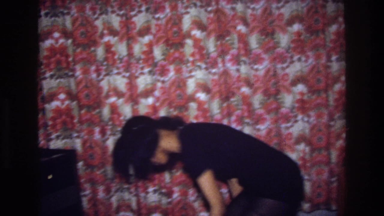 Woman bends low while dancing beside red floral curtains in a dark interior.