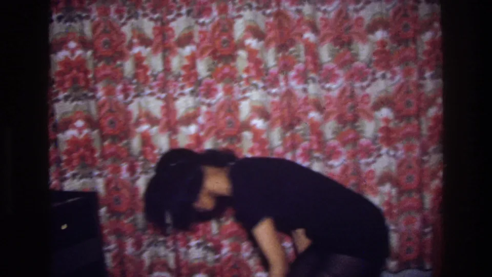 The dancer bends forward in a dim Bronx interior, with red floral curtains filling most of the background.