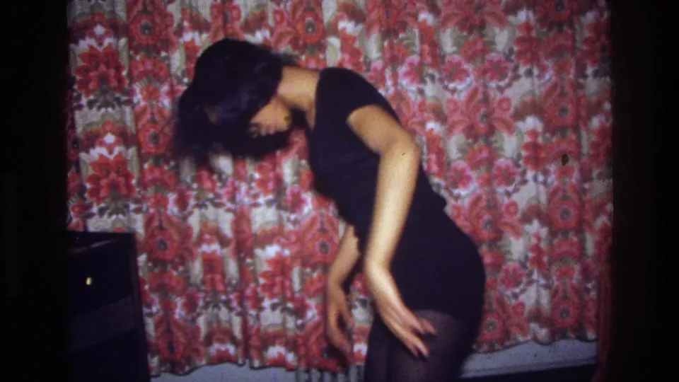 A 1975 home-movie frame shows the woman in profile, arm lowered, dancing in front of red floral curtains.