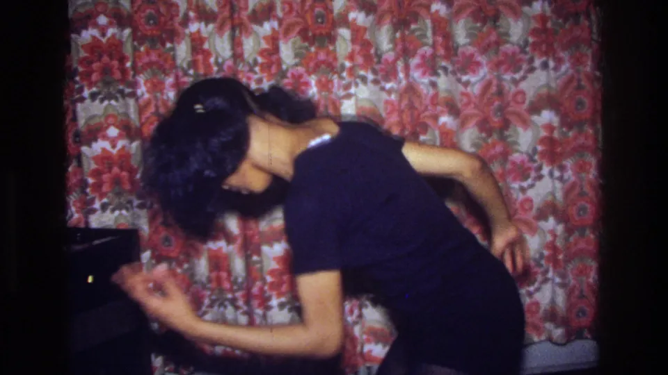 The woman leans into a sideways dance gesture in the Bronx interior, framed by red floral curtains and dim 8mm exposure.