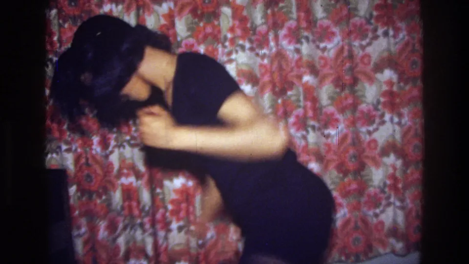 In 1975 Bronx home-movie footage, a young woman in a dark dress bends forward mid-dance before a red and beige floral curtain.