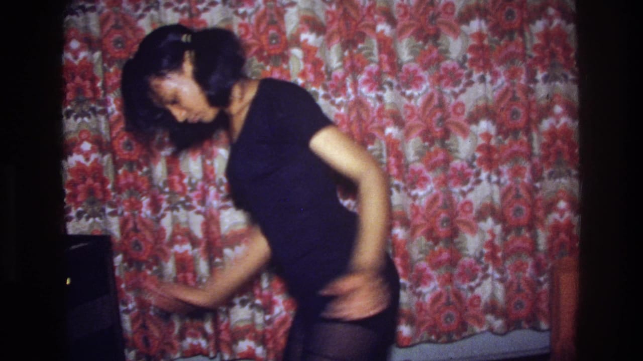 Young woman sways sideways with one arm extended near a floral curtain.