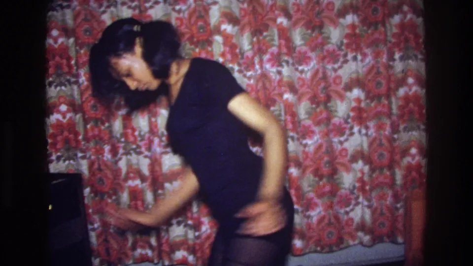 The dancer sways sideways in a dark dress, one arm low and extended, in a Bronx interior documented on 8mm film.