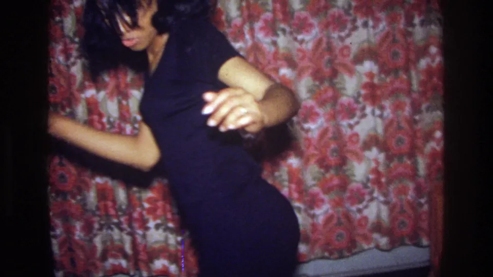 A young woman moves close to the camera with one arm raised, her face partly obscured by hair, in a 1975 Bronx home interior.