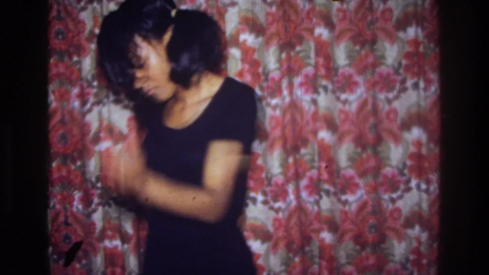The dancer gestures with both arms in front of the floral curtain, captured with soft focus and warm 8mm color in the Bronx.