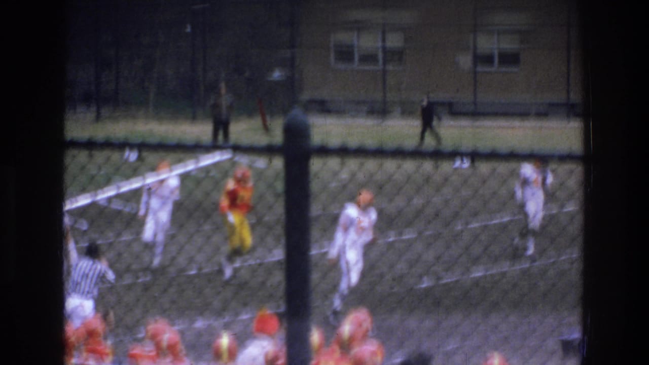 1975 Bronx football game action seen through a chain-link fence, with players in white and orange uniforms on the field.