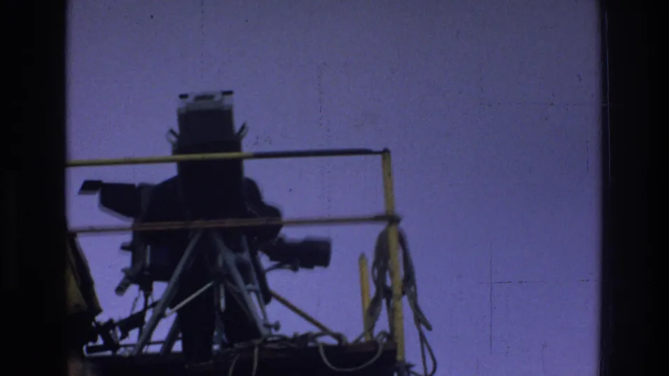 The large camera and tripod remain framed in silhouette, showing the raised platform structure and cables.