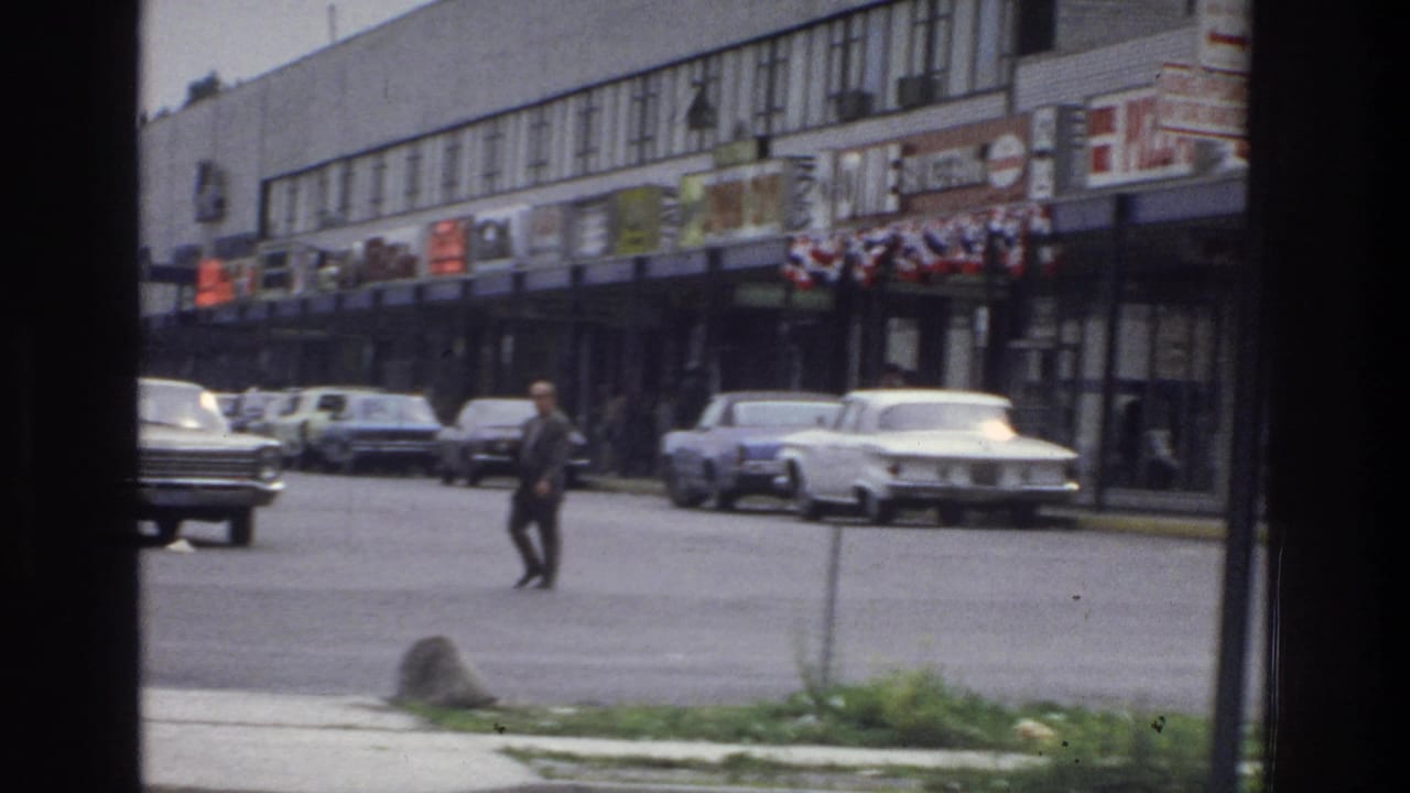 1975 Bronx home-movie frame showing a suited man crossing near parked sedans and storefronts, viewed through a dark frame edge.