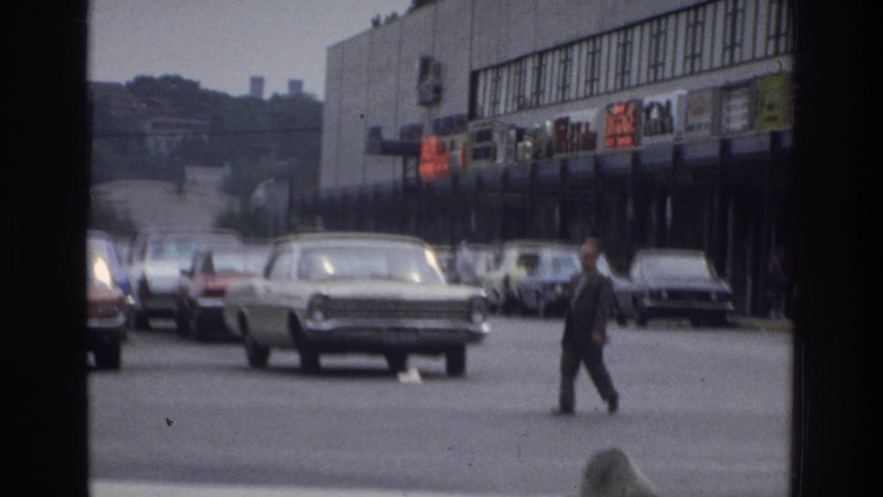 Man in a dark suit walking across a road with parked cars and storefronts behind him.