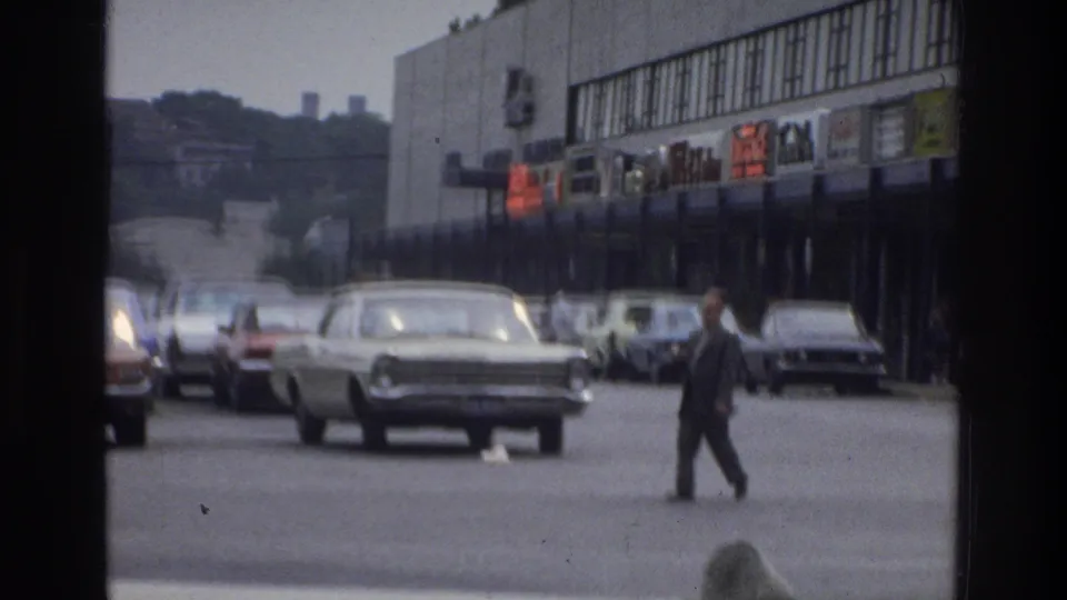1975 Bronx street view with a suited man mid-crossing, several parked cars, storefront signage, and a dark foreground frame.